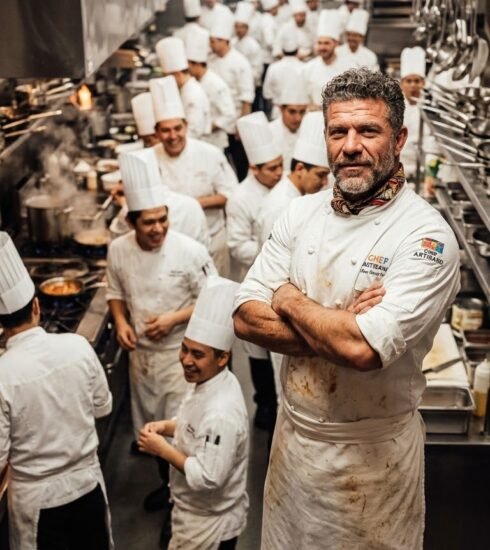 Cocinero maduro de barba gris, brazos cruzados y pañuelo al cuello, en primer plano frente a una cocina abarrotada de cocineros idénticos con gorro alto blanco